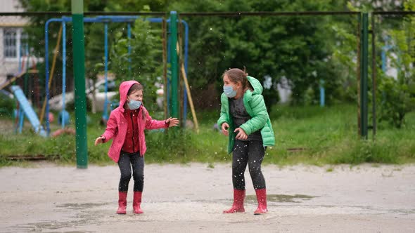 Two little girls in medical masks and rubber boots jump through puddles on a spring day after rain. 