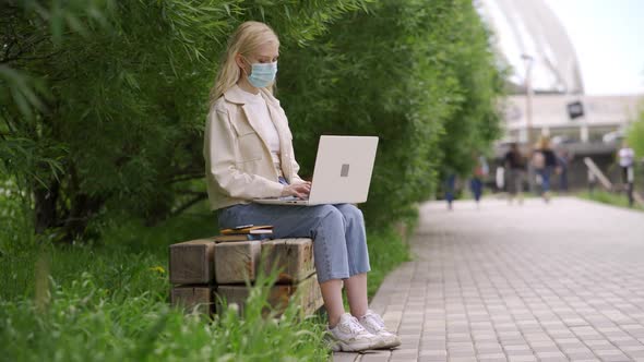 Woman Student in a Medical Mask Makes a Report Using Books and Laptop on a Park Bench