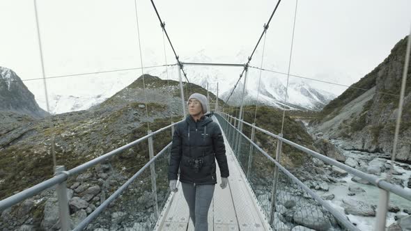 A front on following shot of a woman walking across a swing bridge over a running stream and surroun alt