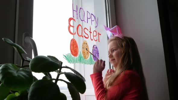 Cute little girl in bunny ears painting Easter eggs with green spring grass on window at home alt