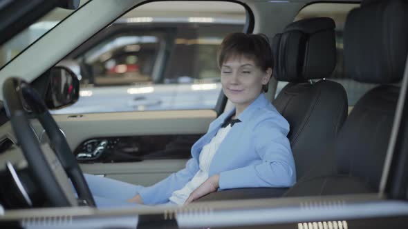 Young Woman with Short Haircut in Formal Wear Blue Suit Sitting Inside a Car in Front Passanger Seat alt