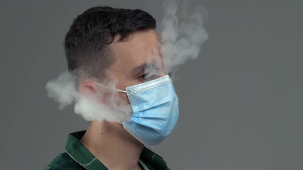 Young Caucasian Male Exhaling Steam From Vape Through Medical Mask