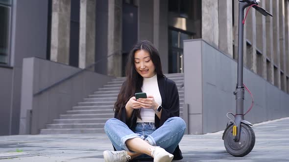 Asian Girl Sitting on the Concrete Ground Near Own e-Scooter on the Beautiful Urban Building alt