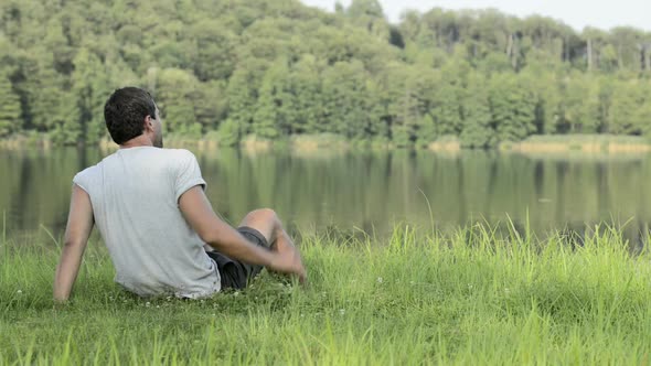 Young Man in Nature Seated on Grass Look Around on Lake Shore in Sunny Summer Day Outdoor