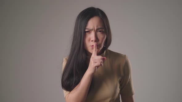 Asian woman putting index finger on lips meaning keeping secret and standing over background.