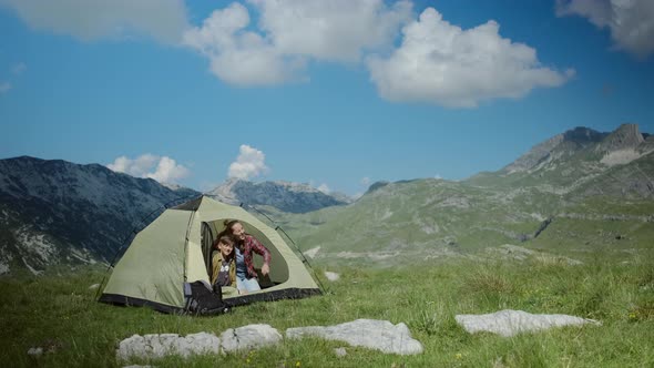 Happy millennial woman in blue t-shirt with her son opening tent and looking at beautiful mountain alt