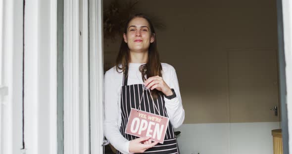 Smiling caucasian waitress standing in door, holding open sign, looking at camera alt
