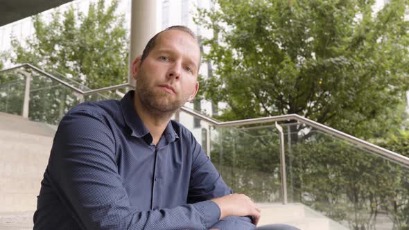 A Caucasian Man Looks Seriously at the Camera As He Sits on a Staircase in an Urban Area