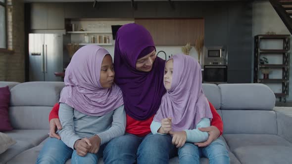 Loving Arab Woman in Hijab Embracing Two Cute Multiethnic Little Daughters on Sofa at Home alt