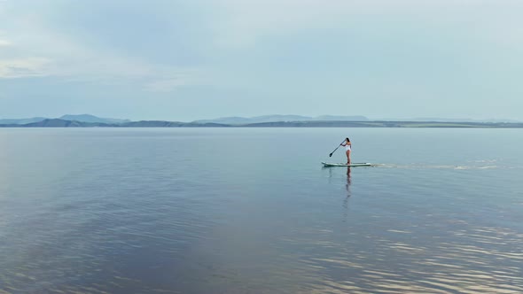 Drone Shot of a Young Woman with Long Hair Floating on a Paddleboard on a Mountain Lake at Sunset