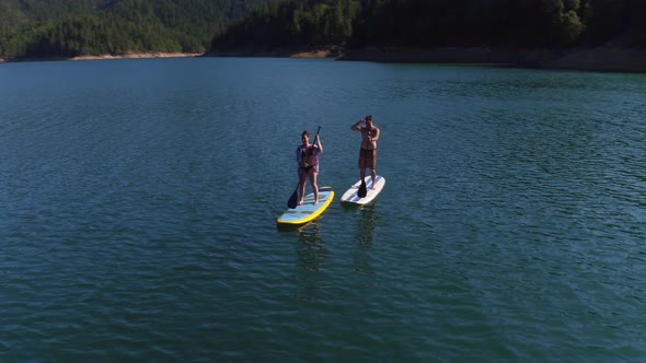 Aerial drone shot of couple paddling stand up paddle boards on lake alt