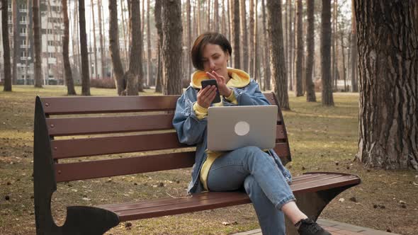 Woman sitting on bench in city park with smartphone in hands, scrolling news alt