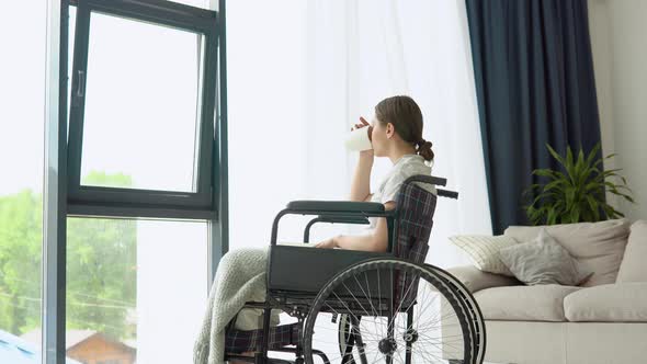 Young Woman Sitting on the Wheelchair Reading a Book and Drinking Coffee at Home alt