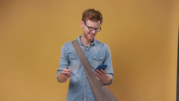 Young Bearded Man Holds Credit Card and Smartphone Isolated on Orange Background alt