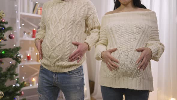 Unrecognizable Adult Caucasian Man and Pregnant Woman Standing Indoors Holding Bellies alt