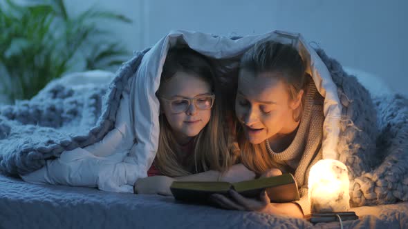 Mom and Daughter Reading Book in Bed Under Blanket alt