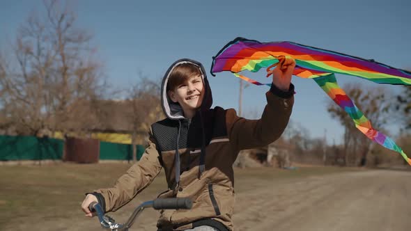 A Happy Boy with a Flying Rainbow Kite Rides a Bicycle on the Road alt
