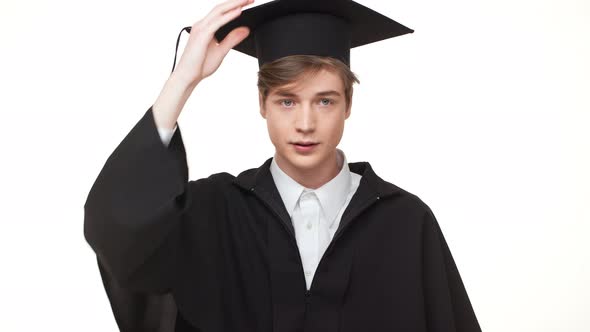 Young Caucasian Graduate in Black Robe Rearranging His Square Academic Cap on White Background alt