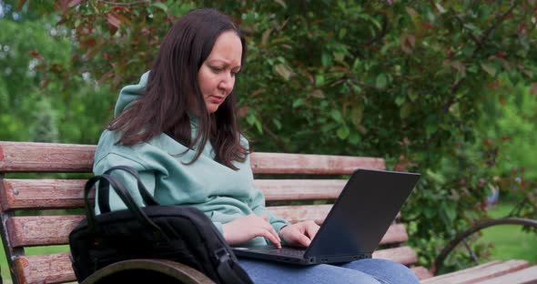 Brunette Woman Freelancer Is Using For Job Laptop Sitting In Park On Bench