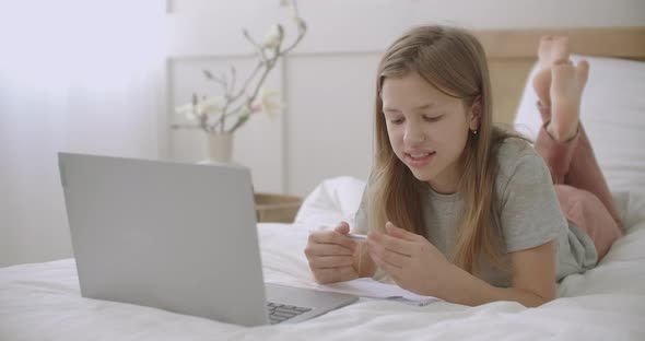 Teen Girl Is Lying on Bed with Laptop and Exercise Book and Talking, Preparing To Doing Homework alt