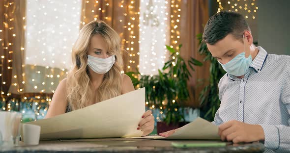 Blonde Woman and Handsome Man in Masks Read Menu at Table alt