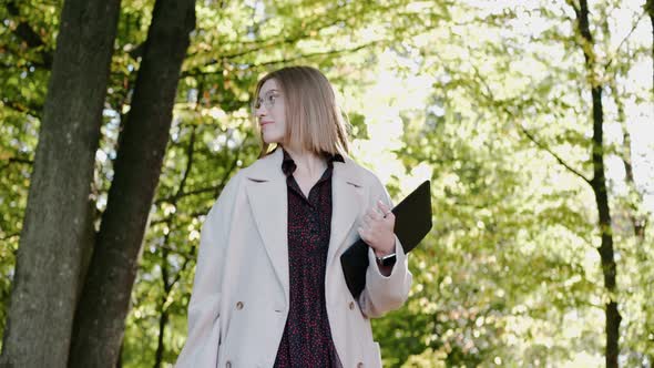 Young Girl Walks in the Park on a Sunny Day Wearing Coat and Drinking From a Cup alt