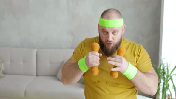 Funny Fat Man in Sportswear Is Boxing with Dumbbells Looking at Camera at Home