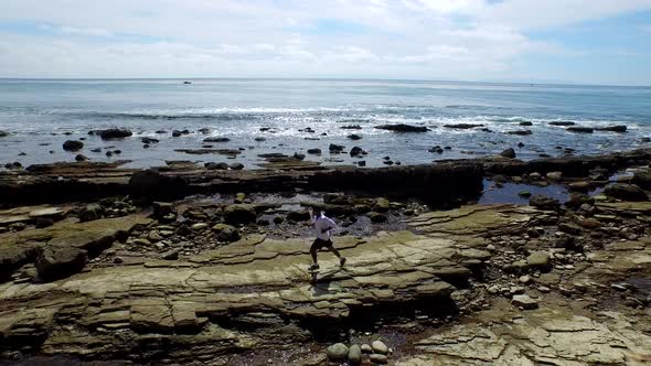 Side view tracking shot of a young man running on a rocky ocean beach shoreline. alt