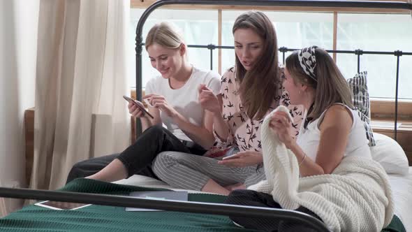 Three Girls sit and chat on a bed in a room, they look at photos on smartphones and smile alt