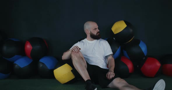 Zoom in Portrait of Young Bearded Athlete Man Sitting on Gym Floor Near Cross Fit Balls Looking at alt