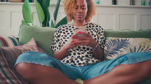 Young Blonde African American Woman Plays Game on Phone Using Mobile Application alt