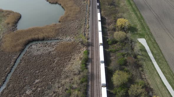 Aerial view of freight train waiting on tracks in rural landscape with retention pond.