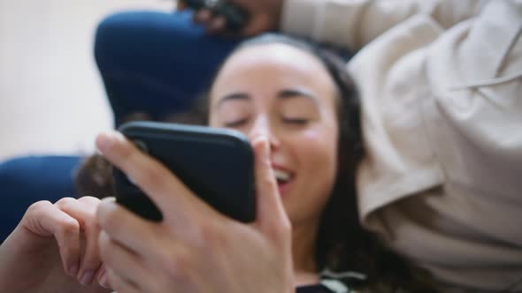 Close Up Of Relaxed Couple At Home On Sofa Watching TV And Checking Social Media On Mobile Phone