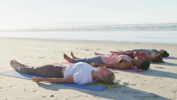 Group of diverse female friends practicing yoga, lying on mats at the beach alt