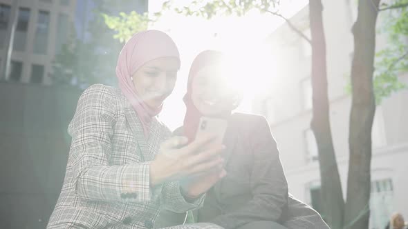 Two Young Muslim Women Wearing Hijab Headscarf Looking at Phone, Sitting Together at Bench Laughing