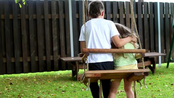 boy and girl ride on a swing in the village. older brother hugging little sister.