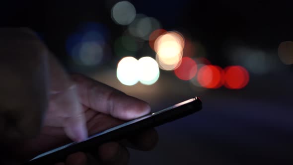 Hand of a Man With a Smartphone in the City at Night Against the Background of Blurry Lights of Cars alt