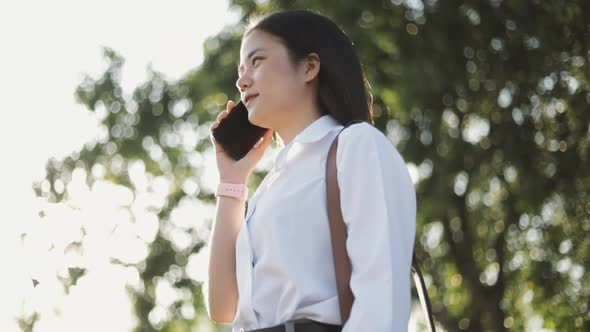 A beautiful Asian female office worker in a white shirt is talking on the phone outside the morning. alt