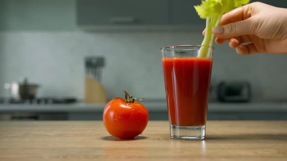 Female hand puts celery stalk into glass with tomato juice. Close up