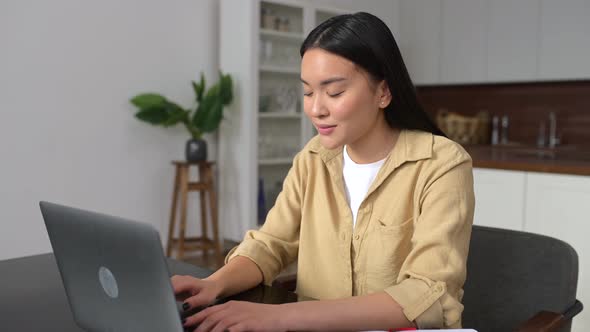 Intelligent Asian Female Looking at Camera Sitting at the Desk in Front of Laptop