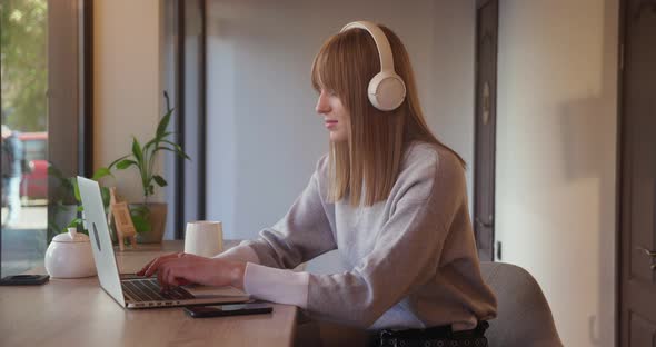 Young and Beautiful Blonde Woman in Headphone Sitting in Coffee Shop is Working on Laptop Computer alt