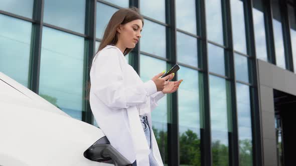 Young Woman is Standing Near the Electric Car and Looks at the Smart Phone alt