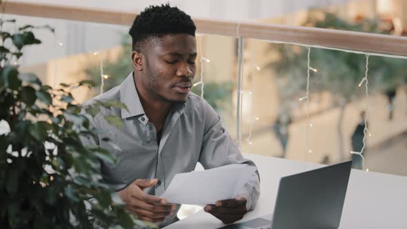 Concentrated Frustrated Upset African American Man Sitting at Desk with Laptop Looking at Documents alt