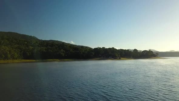Drone footage flying over calm steamy lake (Tinaroo Dam) at sunrise towards a campsite on the bank. alt
