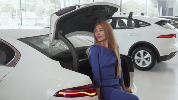 Beautiful Happy Woman Sitting in Open Trunk of Her New Car at Dealership Salon