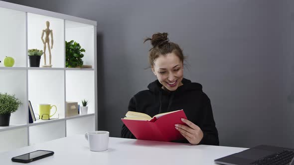 Beautiful Girl in Black Hoody Is Interestedly Reading Book at Home and Smiling