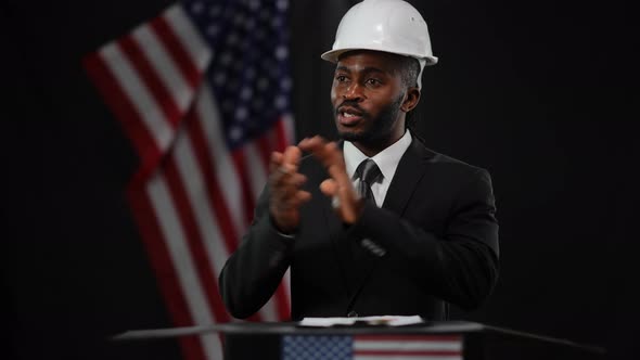 Positive African American Man in Suit and White Hard Hat Talking Gesturing and Smiling Presenting alt
