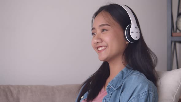 Attractive young Asian woman listening to music on headphones While sitting sofa in the living room.