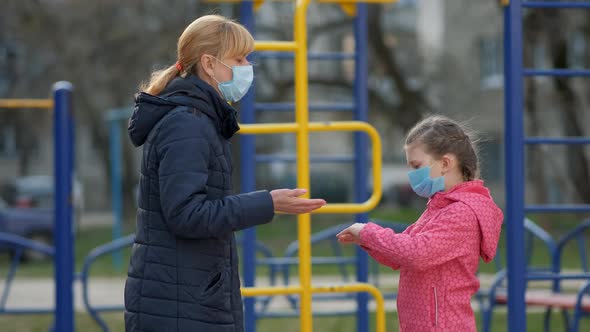 A Girl in a Medical Mask. Her Mother Sprays Disinfectant on Her Hands in the Playground. The Concept