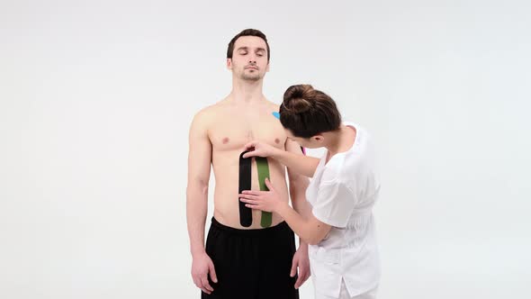 Female therapist applying kinesiology tape on a man's abdomen on the light background.  alt